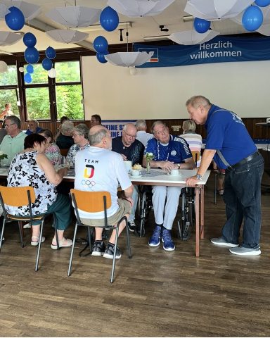 Eine Gruppe von Menschen sitzt an Tischen und unterhält sich, umgeben von blauen Ballons.