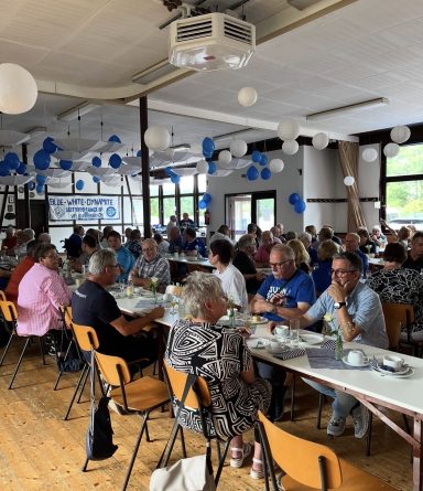 Menschen sitzen an langen Tischen in einem hellen Raum mit blauen und weißen Dekorationen.