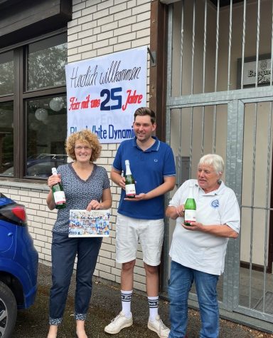 Drei Personen stehen vor einem Gebäude und halten Flaschen für ein Jubiläum.